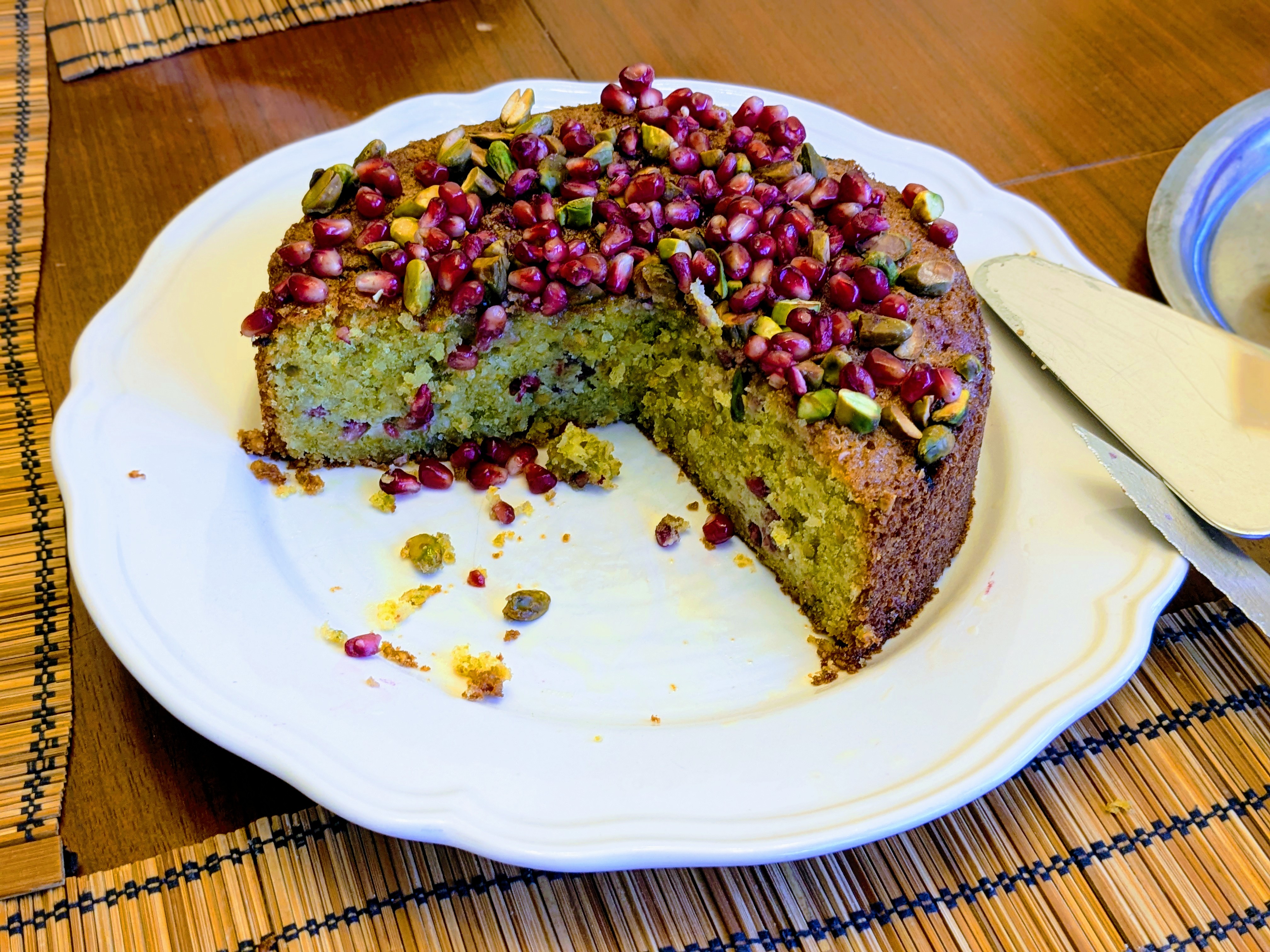 Un gâteau à la pistache et à la grenade sur une assiette blanche. Le bord est brun, l'intérieur vert, et le gâteau est parsemé de graines de grenade et de pistaches grossièrement hachées.